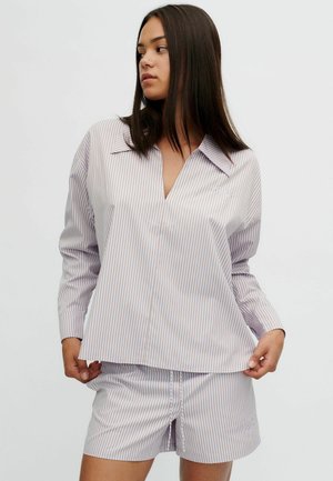 Femme aux longs cheveux foncés portant une chemise ample rayée légère assortie et un short, debout et regardant sur le côté sur un fond uni.