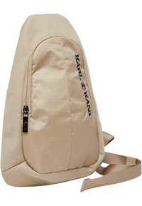 Beżowy plecak o teksturowanym wykończeniu, z przednią kieszenią na zamek i bocznym logo. Kształt jest lekko zakrzywiony, z pojedynczym regulowanym paskiem.