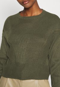 Olijfgroene chunky gebreide trui met een ronde halslijn, ribbelrand en textuurpatroon details. Het materiaal lijkt zacht en comfortabel.