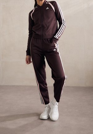 Osoba nosząca ciemnobrązowy dres Adidas z białymi paskami i białe tenisówki, stojąca na tle jednolitej jasnej ściany.
