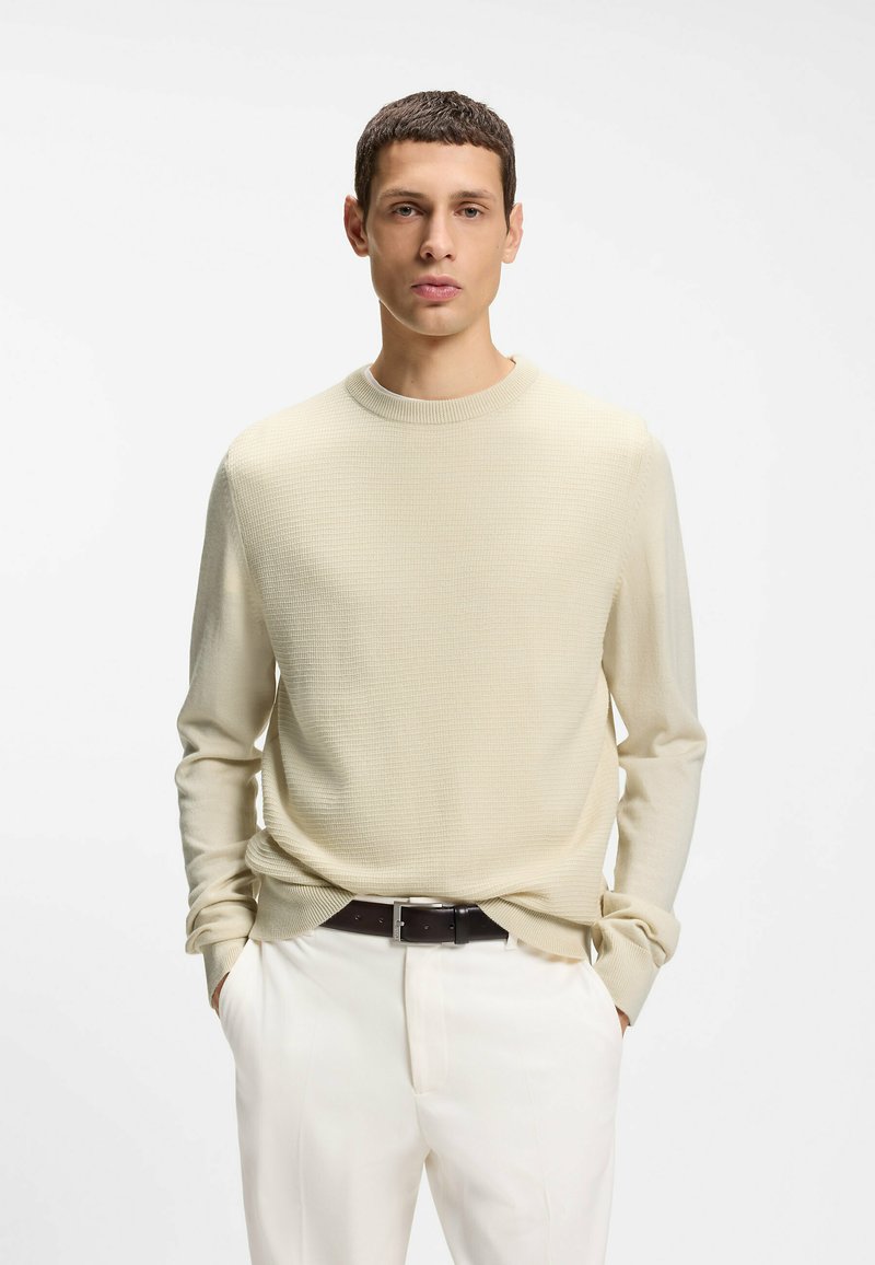 Pull en maille beige clair avec un motif texturé, col rond et manches longues, associé à un pantalon blanc et une ceinture foncée.