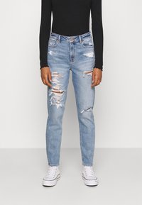 Světle modré džínové kalhoty s opotřebovaným vzhledem, mnoha roztrženinami, vysokým pasem a zúženými nohavicemi, v kombinaci s černým topem s dlouhým rukávem a bílými teniskami.