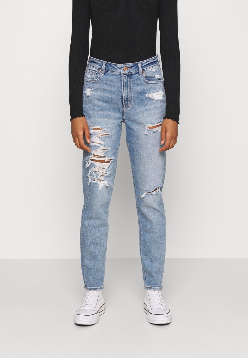 Světle modré džínové kalhoty s opotřebovaným vzhledem, mnoha roztrženinami, vysokým pasem a zúženými nohavicemi, v kombinaci s černým topem s dlouhým rukávem a bílými teniskami.