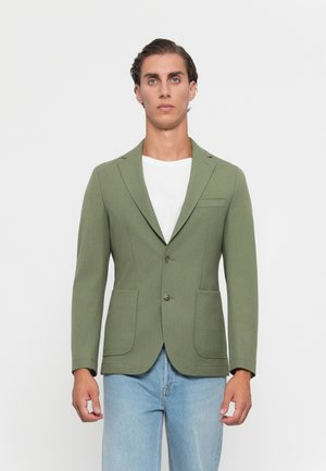 Young man wearing a light olive green blazer over a white T-shirt and light blue jeans, standing against a plain white background.