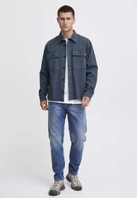 Temno modra srajčna jakna z dvema sprednjima žepoma, v kombinaciji z belo majico, svetlo modrimi jeans hlačami in sivimi pohodniškimi čevlji. Minimalističen dizajn.