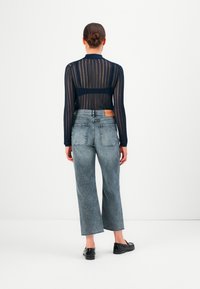 Femme avec les cheveux en chignon portant un top long à manches longues en voile bleu marine, un jean gris taille haute et cropped, et des mocassins noirs, se tenant de dos.