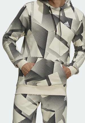 Homme portant un sweat à capuche beige et un pantalon avec des motifs géométriques noirs et des logos Adidas, les mains dans les poches du sweat.