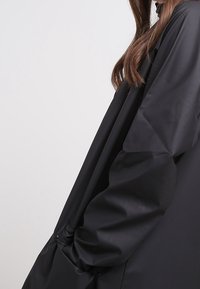 Svart vattentät kappa med långa ärmar, avslappnad passform, slät textur och knappdetaljer nära fållen. Minimalistisk design, utan mönster.