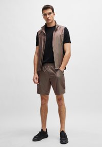 Chaleco ligero y brillante de color marrón con un cuello alto, combinado con pantalones cortos a juego. Una camiseta negra debajo y zapatillas deportivas negras completan el conjunto.