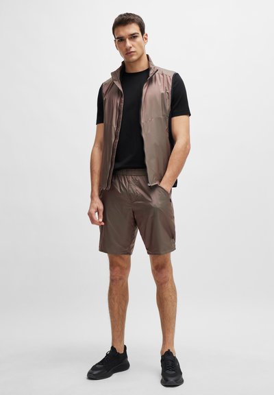 Gilet léger et brillant de couleur brune avec un col haut, associé à un short assorti. Un t-shirt noir en dessous et des chaussures de sport noires complètent le look.