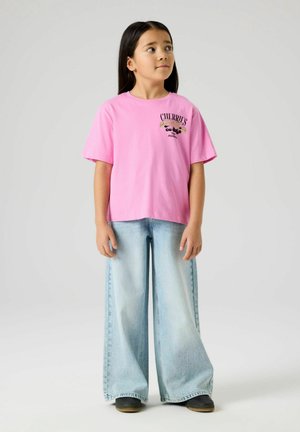 Dziewczyna o długich ciemnych włosach, ubrana w różowy T-shirt z wzorem czereśni oraz szerokie jasnoniebieskie dżinsy, stojąca na jednolitym tle.