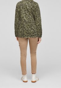 Een persoon in een groene camouflagejas, beige slim fit cargobroek en witte hoge sneakers, staande tegen een effen grijze achtergrond.