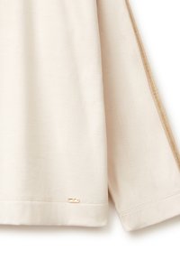 Krémově zbarvená mikina s měkkou texturou, zdobená zlatým kovovým doplňkem na lemu a jemným bočním designovým detailem.