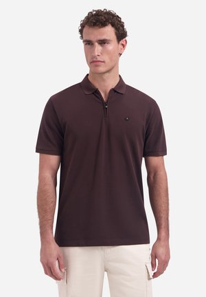 Man die een donkerbruin poloshirt met korte mouwen en een klein embleem draagt, gecombineerd met lichtbeige cargobroeken, staand tegen een effen achtergrond.