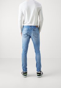 Hombre de pie de espaldas, con jeans slim de color azul claro, camisa blanca de manga larga y zapatillas negro y blanco con detalles verdes.