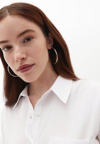 Camicia bianca con colletto e taschino sul petto. Il modello indossa grandi orecchini a cerchio argentati. Stile minimalista e texture liscia.
