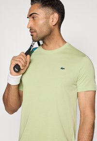 Ανοιχτό πράσινο κοντομάνικο μπλουζάκι με στρογγυλή λαιμόκοψη και μικρό κεντημένο λογότυπο. Μαύρη ρακέτα τένις κρατιέται από άτομο που φοράει βραχιολάκι στον καρπό.