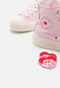 Różowe trampki wysokie Converse z detalem serca na łatce logo oraz dwoma czerwonymi akcesoriami w kształcie serca z białymi wzorami róż w pobliżu.