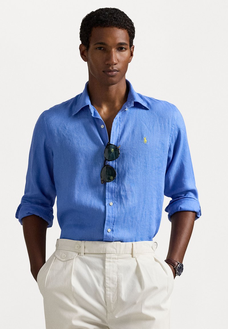 Lyseblå linneskjorte med krage som kan kneppes ned, rullede ermer og et lite gult logo. Matcher med kremfargede, pregede bukser.