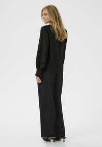 Schwarze leichte Bluse mit strukturiertem Stoff, langen Ärmeln und lässiger Passform, kombiniert mit weiten schwarzen Hosen und spitzen High Heels.