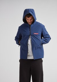 Veste à capuche bleue avec zip frontal, dotée d'une poche poitrine horizontale avec un logo rouge, en matériau léger et avec deux poches latérales.