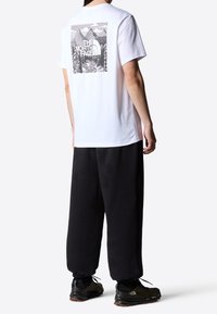 Person wearing white T-shirt with black mountain graphic and "The North Face" text, black pants, and dark sneakers standing sideways.