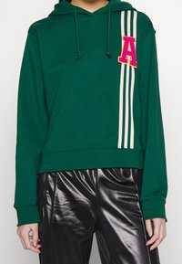 Felpa verde con tasca frontale, coulisse, strisce bianche sulle maniche e una toppa rosa con la lettera "A" sul petto. Pantaloni neri lucidi sotto.