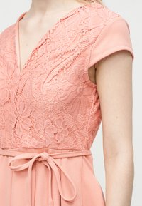 Žena v ružových šatách s kvetinovou čipkovanou časťou na živote, krátkymi rukávmi, výstrihom do V a látkovým opaskom previazaným v páse.