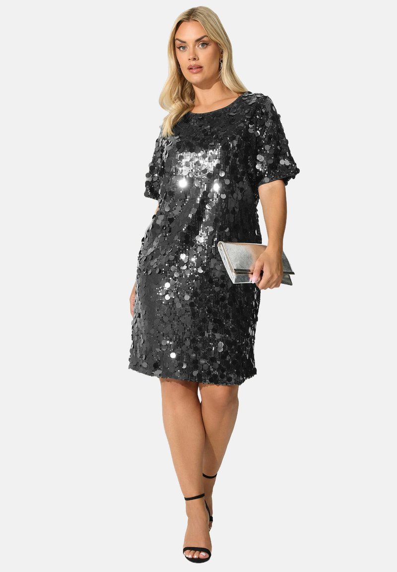 Schwarzes, mit Pailletten besetztes Shiftkleid mit kurzen Ärmeln, runden Ausschnitt und lockerer Passform. Das Model hält eine silberne Clutch und trägt dazu Riemchensandalen.
