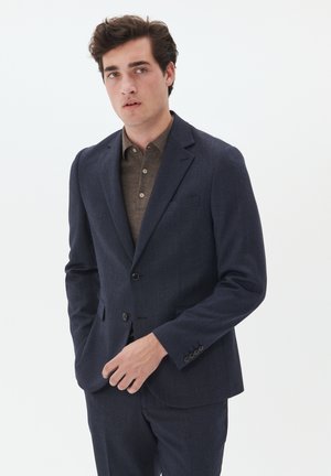 Marineblaues Blazer aus strukturiertem Stoff, einreihige Verarbeitung, Reverskragen und Knöpfe. Über einem braunen Hemd mit Kragen getragen.