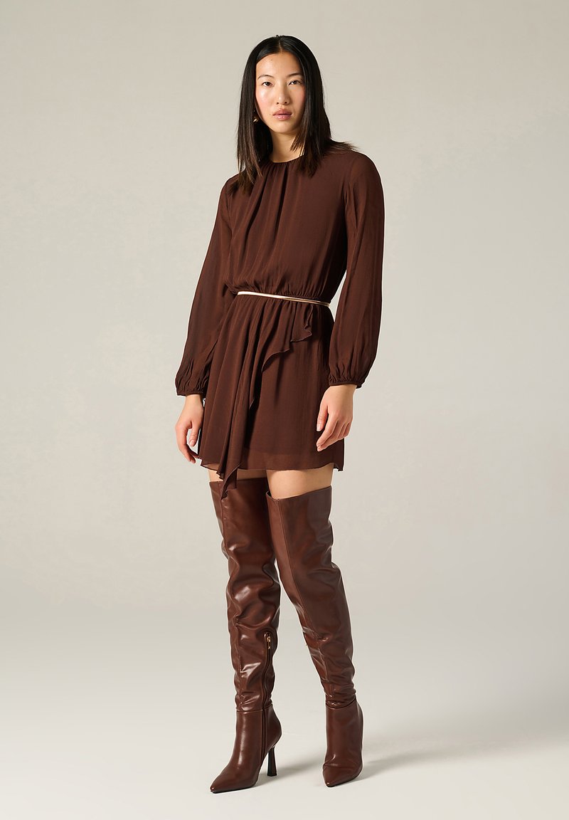 Robe longue marron à manches longues avec une taille cintrée et un ourlet à volants, portée avec des bottes en cuir marron au-dessus du genou et une fine ceinture blanche.