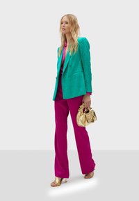 Blazer texturizado turquesa, blusa fucsia y pantalones fucsia de pierna ancha. Sosteniendo un bolso dorado brillante y usando sandalias de tacón doradas.