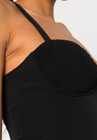 Svart klänning med en ribbad bål, med tunna axelband. Texturen är slät och framhäver den strukturerade formen på toppen.