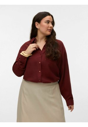 Femme aux longs cheveux foncés portant une chemise bordeaux boutonnée et une jupe beige, ajustant le col de la main gauche, regardant vers la droite.