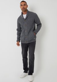 Szary sweter kardigan z dzianiny z kołnierzem szalowym, ozdobiony drewnianymi guzikami, noszony na białej koszulce, zestawiony z ciemnymi dżinsami i białymi trampkami.