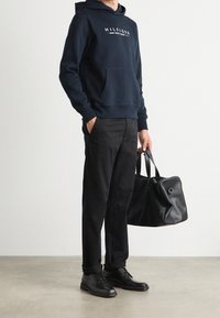 Námořnická mikina s kapucí a přední kapsou, černé kalhoty a černé kožené boty. Drží černou koženou tašku s zakřiveným tvarem a kovovými detaily.