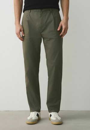 Mężczyzna noszący oliwkowozielone spodnie o prostych nogawkach z przednimi zakładkami oraz białe tenisówki z czarnymi i szarymi detalami.