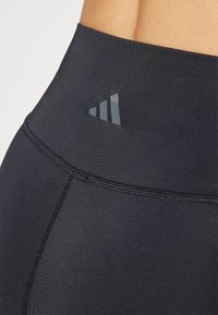 Czarne legginsy z wysokim stanem, o eleganckim designie, gładkiej fakturze oraz subtelnym, błyszczącym logo z wzorem trzech pasków w okolicy pasa.