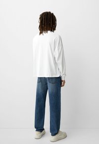 Camicia bianca a maniche lunghe con una vestibilità rilassata, abbinata a jeans baggy blu chiaro e sneakers beige. Design semplice, texture del tessuto liscia.