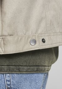 Béžová semišová bunda se stříbrnými detaily, s patentním knoflíkem a prošíváním na lemu. Nahoře je vrstvena nad olivově zeleným svetrem.