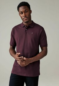 Burgundská polo košeľa s krátkymi rukávmi, golierom, tromi gombíkmi a malým logom na ľavej hrudi. Hladká tkanina a prispôsobený strih.