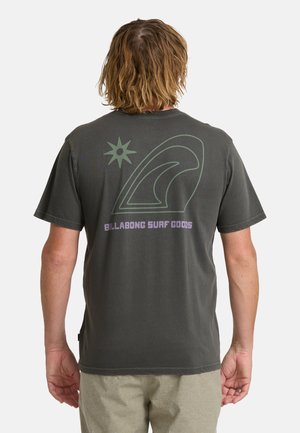 Persona con capelli di media lunghezza che indossa una maglietta scura con grafica di onda e sole e testo "Billabong Surf Goods" sulla schiena.