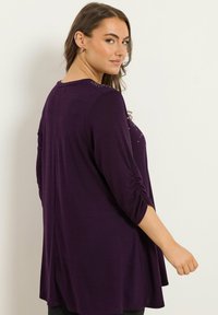 Lila långärmad topp med rynkade ärmar och dekorativa nitar på axeln. Utställd design ger en avslappnad passform och en flödande silhuett.