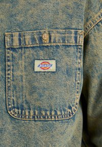 Giacca di jeans con una finitura blu sbiadita, dotata di una tasca sul petto sinistro chiusa da un bottone di metallo e un colorato patch del logo "Dickies".