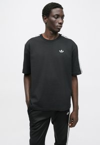 Μαύρο βαμβακερό κοντομάνικο μπλουζάκι με στρογγυλή λαιμόκοψη, με λευκό λογότυπο στο στήθος και λευκές ρίγες κατά μήκος των ώμων.