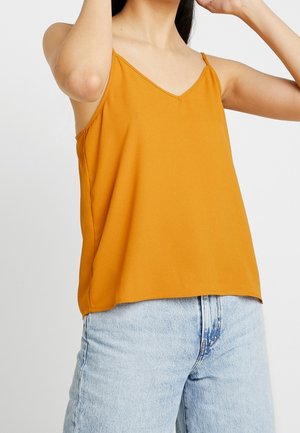 Woman wearing a mustard yellow sleeveless top and high-waisted light blue denim jeans against a plain background.