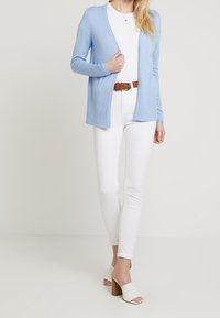 Hellblauer Cardigan aus weichem Stoff, offenes Frontdesign, getragen über einem weißen Oberteil, kombiniert mit weißen Jeans und gemusterten weißen Blockabsatz-Mules.