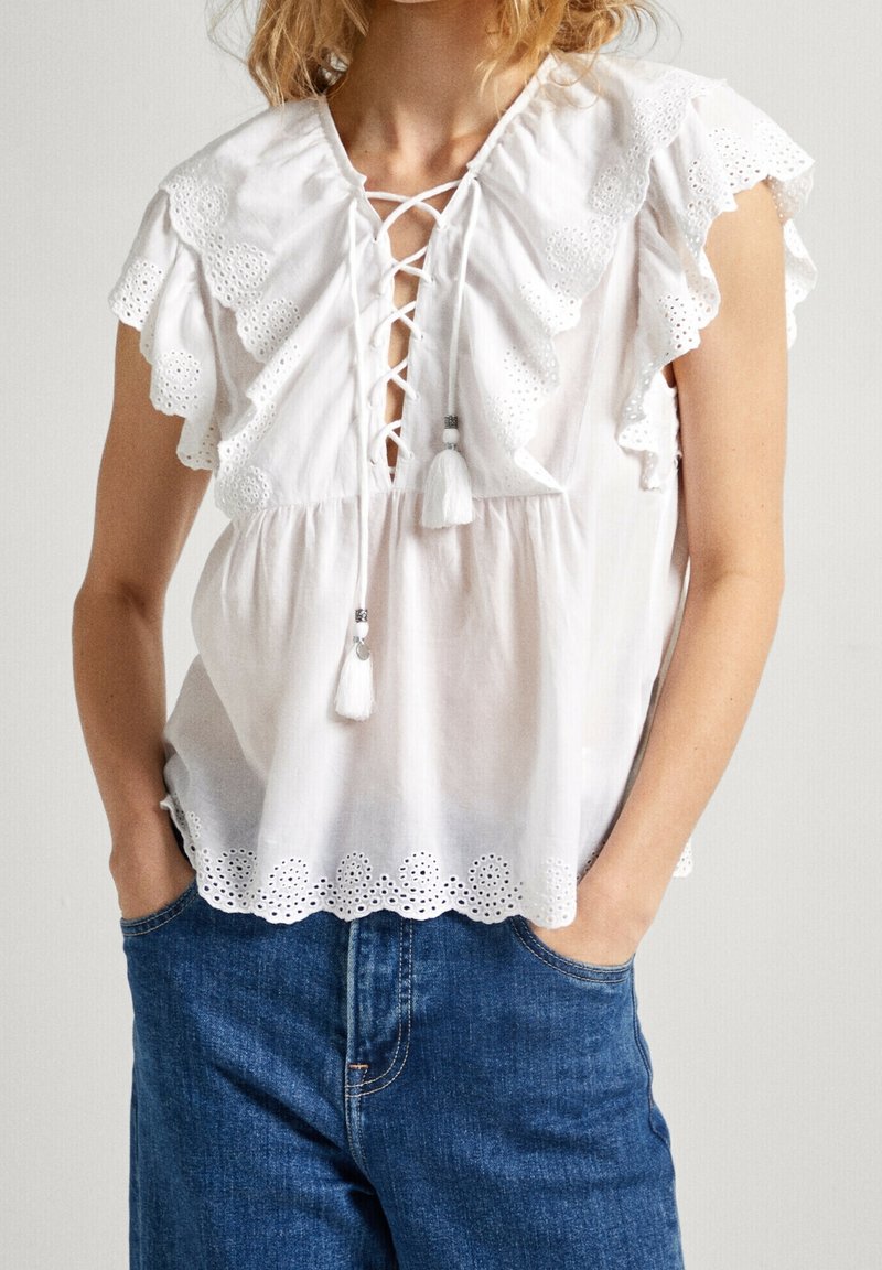 Hvid bluse med volangærmer, snøre foran og frynse-detaljer. Udsmykket med hulbroderi og bølget kant. Kombineret med blå jeans.