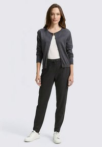 Tamnosivi kardigan s okruglim izrezom na vrat, zatvaranje na gumbe; uparen s crnim hlačama s tankim prugama i bijelim tenisicama. Glatka tekstura, ležeran dizajn.