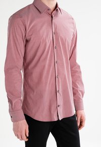 Chemise boutonnée rayée rouge et blanche avec un col, des manches longues et des boutons noirs. Tissu lisse, design ajusté et détails aux poignets.
